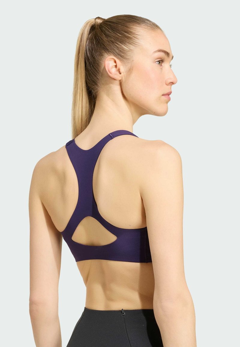 Femme aux cheveux blonds en queue de cheval portant un soutien-gorge de sport dos nageur violet foncé et un legging noir taille haute, face à droite avec le dos visible.