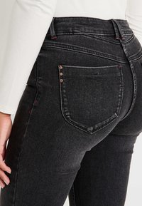 Primer plano de una persona que lleva unos jeans de mezclilla negros de cintura alta con detalles en los bolsillos traseros, combinados con una blusa blanca de manga larga.