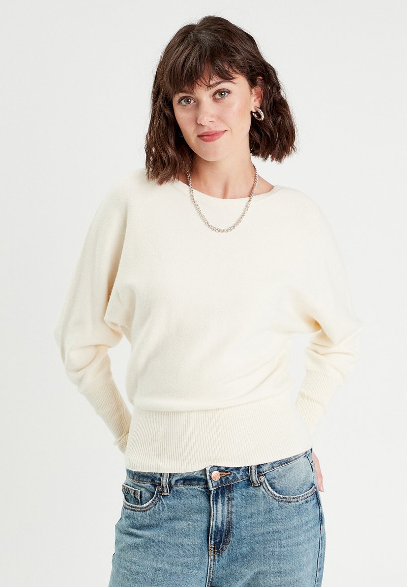 Cremefarvet sweater med et løst, draperet design, ribbet nederste kant og manchetter, parret med højtaljede blå jeans. Enkel sølvfarvet kædehalskæde.
