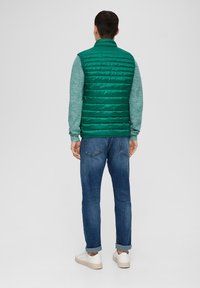 Grüne Steppweste mit quiltierter Textur, hohem Kragen, getragen über einem grauen Pullover, kombiniert mit blauen Jeans und weißen Turnschuhen.