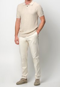 Polo de punto beige con mangas cortas y cierre de tres botones, combinado con pantalones de lino blanco roto y zapatillas de cordones de un beige claro.