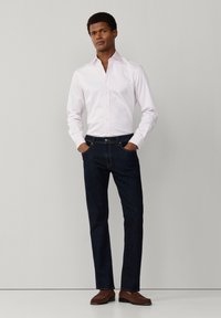 Muž stojí s rukami v kapsách, oblečený v bielej košeli, tmavomodrých džínsoch a hnedých topánkach, na jednoduchom svetlosivom pozadí.