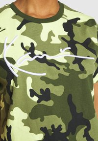 Zielona koszulka w kamuflażu z okrągłym dekoltem i krótkimi rękawami. Posiada białe haftowane napisy na klatce piersiowej. Miękka bawełniana tekstura.