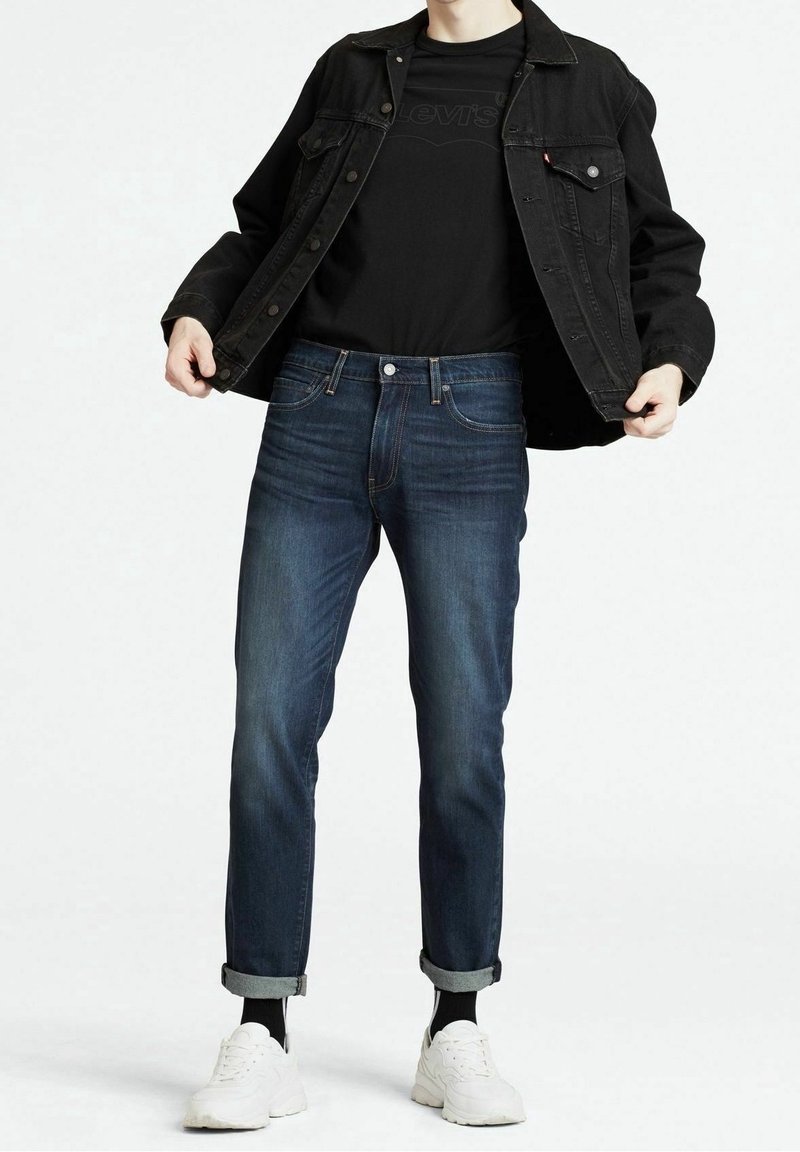Chaqueta de denim negra sobre una camiseta negra, combinada con jeans azul oscuro enrollados en los puños y zapatillas blancas. Estilo minimalista, líneas limpias.