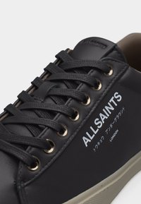 Μαύρο δερμάτινο αθλητικό παπούτσι με μαύρα κορδόνια, χρυσά μεταλλικά τρύπες και την επωνυμία "ALLSAINTS" με ιαπωνικό κείμενο και τη λέξη "LONDON" στο πλάι.