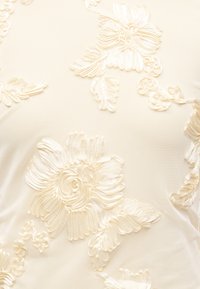 Tissu crème avec broderie florale en relief et texturée comportant de grandes fleurs et des feuilles dans un motif délicat.
