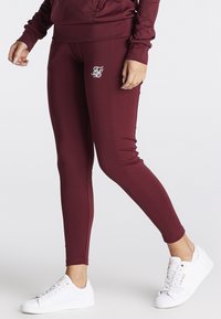 Maroone leggings lavet af strækbart stof med en tætsiddende design, der har en hvid logoaccent på hoften. Båret med hvide sneakers.