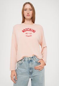 Haut rose clair à manches longues avec broderie rouge "Moschino 86-23 Jeans". Porté avec un jean en denim bleu clair taille haute.