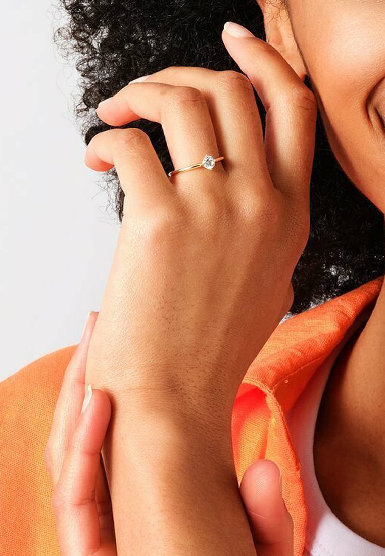 Anello d'oro con una pietra preziosa rotonda e trasparente, indossato su una mano con tonalità di pelle media. La mano tocca delicatamente il polso, con uno sfondo testurizzato.