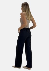 Zwarte wijde broek met een elastische tailleband, gecombineerd met een beige geribde tanktop; model staat met de rug naar voren, handen in de broekzakken.