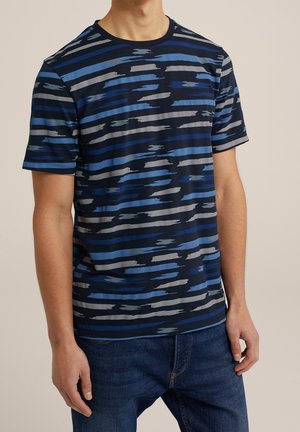 Homme portant un t-shirt bleu marine à manches courtes avec des rayures horizontales irrégulières dans des nuances de bleu et de gris, associé à un jean bleu foncé.