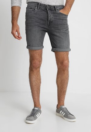 Mand iført opkrogede mørkegrå denimshorts, grå og hvide Adidas sneakers og en langærmet, stribet skjorte med den ene hånd i lommen.