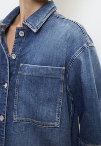Jasnoniebieska kurtka dżinsowa z kołnierzykiem, z przodu zapinana na guziki oraz z kieszenią na piersi. Ma wyblakłą teksturę i kontrastowe szwy.