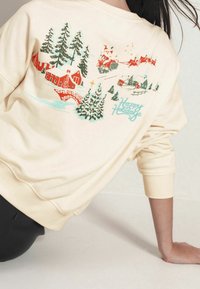 Sweat-shirt couleur crème avec un imprimé festif comprenant des maisons rouges, des sapins verts et le Père Noël. Le texte indique « Joyeuses fêtes ». Coupe décontractée.