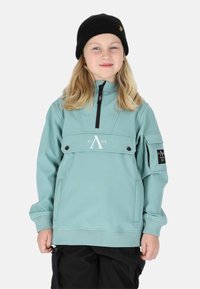 Hellblauer Pullover-Jacke mit hohem Kragen, Vorderasche und schwarzen Akzenten. Mit Reißverschluss und gerippten Bündchen. Schwarze Mütze getragen.