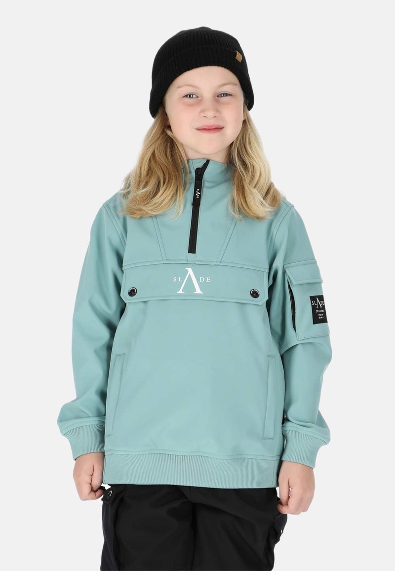 Lys teal pullover jakke med høj krave, forbagslomme og sorte detaljer. Har lynlåslukning og ribbede manchetter. Sort beanie bæres.