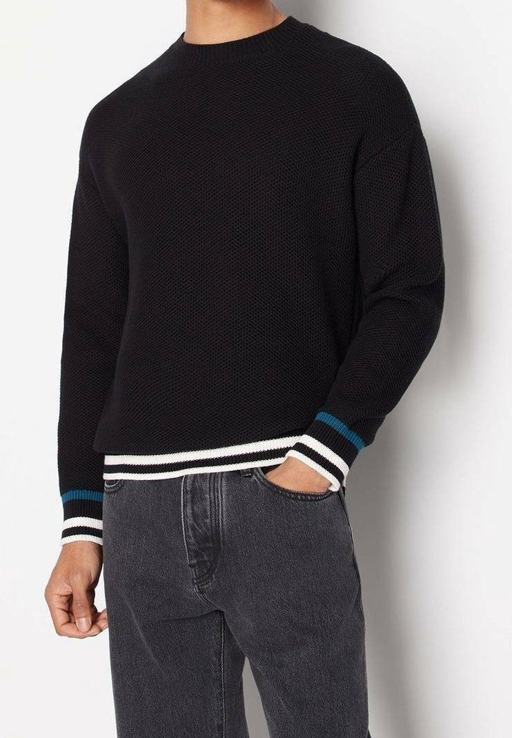 Homme portant un pull noir texturé avec des poignets et un bas rayés blanc, noir et bleu, associé à un jean gris foncé, main dans la poche.