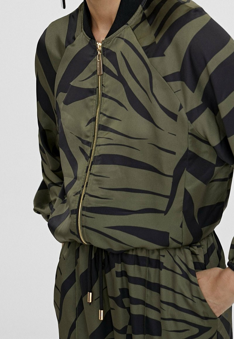 Olivově zelená bomber bunda s černými abstraktními pruhy. Disponuje zipovým zapínáním, žebrovaným límcem a elastickým pasem se šňůrkami.
