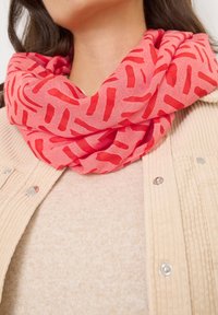 Elegante sciarpa leggera color corallo con motivi astratti rossi, avvolta delicatamente attorno al collo, abbinata a un maglione beige strutturato e a una giacca cordata.