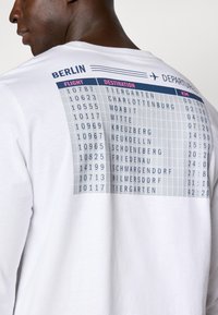 White long-sleeve shirt featuring a printed departure board design on the back, listing flight numbers, destinations, and distances in kilometers.