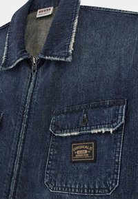 Giubbotto di denim blu scuro, con chiusura a zip, colletto a punta e due tasche sul petto con patella a bottone; include un'etichetta del marchio.