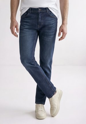 Homme portant un jean bleu foncé et des baskets blanches, debout avec les jambes croisées devant un fond blanc uni.