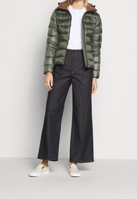 Pufová bunda v tmavě zelené barvě se zipem vpředu a hnědou kapucí, spárována se širokými tmavými denimovými kalhotami a bíl�ými slip-on botami.