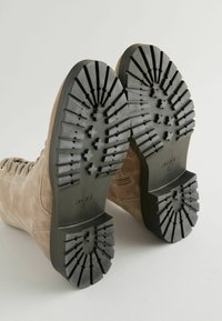 Botas de ante marrón hasta el tobillo con suelas de goma negra, con un tipo de dibujo en la suela para mayor agarre y detalles de cordones a los lados.
