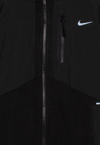 Svart jakke med glatt stoff, fremtredende glidelås foran, brystlomme og blå Nike-logo. Teksturerte svarte paneldetaljer hever designet.