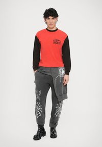 Camisa de manga larga roja y negra con el logo "Aries". Pantalones deportivos grises con gráficos de telarañas blancas y texto. Botines negros hasta el tobillo.