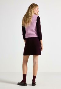 Gilet rosa lavorato a maglia con orlo a costine indossato sopra un vestito bordeaux. La modella indossa calze bordeaux e mocassini neri lucidi. Sfondo neutro.