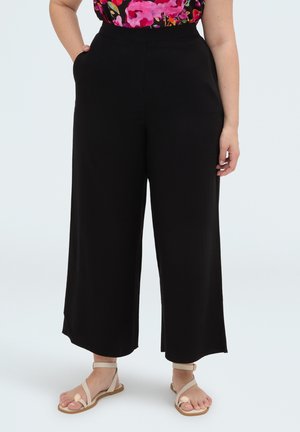 Vrouw die zwarte, wijde cropped broek, een bloeiend top en beige sandalen met open tenen draagt, staat met één hand in haar zak tegen een witte achtergrond.