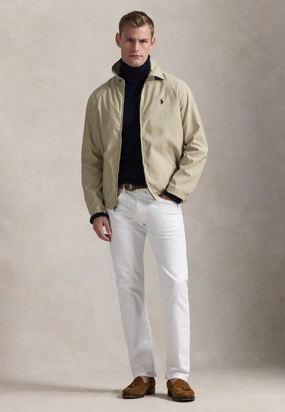 Mand iført beige lynlåstrøje over navy rullekrave, hvide bukser og brune loafers, stående med den ene hånd i lommen mod ensfarvet baggrund.