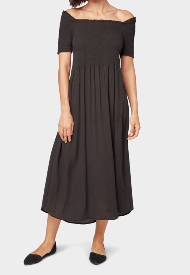 Femme portant une robe midi noire épaules dénudées avec manches courtes et chaussures plates noires, debout devant un fond uni.