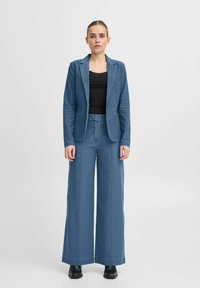 Ljusblå denimdräkt bestående av en skräddarsydd kavaj och vida byxor, kombinerad med en svart topp. Har en enda kavajknapp.