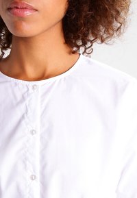 White button-up shirt with a round neckline, featuring a button placket and smooth texture. Fabric appears lightweight and wrinkle-resistant.