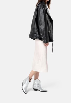 Femme portant une veste en cuir noire sur une jupe midi blanche et des bottines argentées brillantes, debout devant un fond uni.