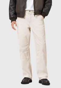Beige corduroy broeken met een recht-pijp ontwerp, voorzien van textuurvariaties en zijzakken, gedragen met een bruine bomberjack en zwarte schoenen.