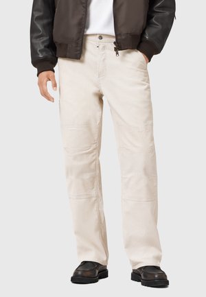 Pantalon en velours côtelé beige avec une coupe droite, présentant des variations de texture et des poches latérales, porté avec une veste bomber marron et des chaussures noires.