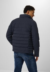 Mörkblå pufferjacka med horisontellt kviltad mönster, hög krage och enhetligt tyg. Bärs med jeans i blått, sedd bakifrån.