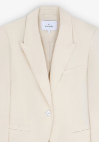 Beige bleiser valmistettu kevyestä kankaasta, jossa on yksittäinen kiiltävä nappi, leikatut läpät ja hienovarainen taskudetaili. Brändilogo näkyvissä sisällä.