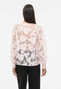 Femme aux longs cheveux noirs portant un chemisier rose transparent à motifs floraux et un pantalon noir, vue de dos sur un fond blanc.