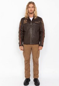 Veste en cuir marron avec des écussons brodés, fermeture éclair à l'avant et accents à rayures rouges. Associée à un pantalon cargo beige et des bottes noires.