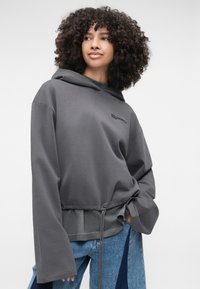 Γκρι φούτερ με κορδόνι στο τελείωμα, μακριά μανίκια και διακριτό λογότυπο. Απαλή ύφανση με χαλαρή εφαρμογή. Φοριέται με μπλε τζην με πα patchwork.