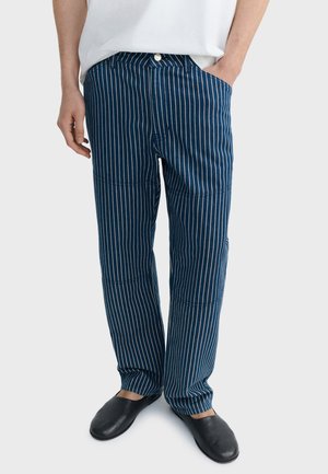 Personne portant un pantalon bleu à rayures verticales blanches, une chemise blanche et des chaussures noires sans lacets, debout avec une main dans la poche.