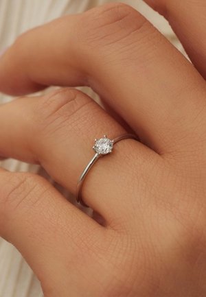 Bague en argent avec une pierre précieuse transparente ronde sertie dans un chaton à six griffes, portée sur une main à la peau texturée. Design simple et élégant.