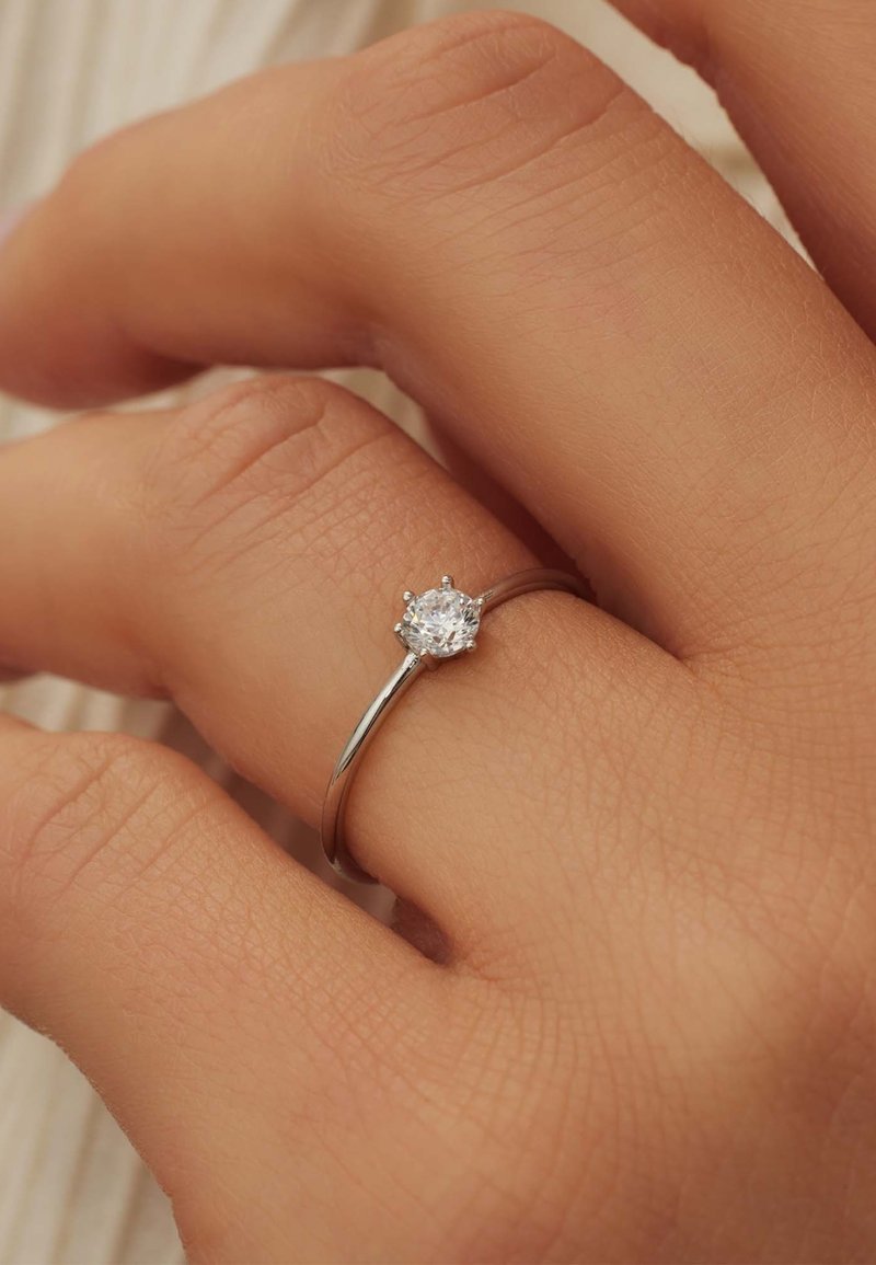 Bague en argent avec une pierre précieuse transparente ronde sertie dans un chaton à six griffes, portée sur une main à la peau texturée. Design simple et élégant.