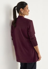Vrouw met donker haar in een paardenstaart, gekleed in een langetermijn bordeauxrode trui en zwarte broek, glimlachend en gericht op een witte muur.