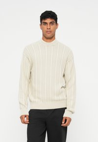 Pullover de malha em bege claro, com riscas verticais e um pescoço alto, apresentando um padrão texturizado e punhos e barra canelados.