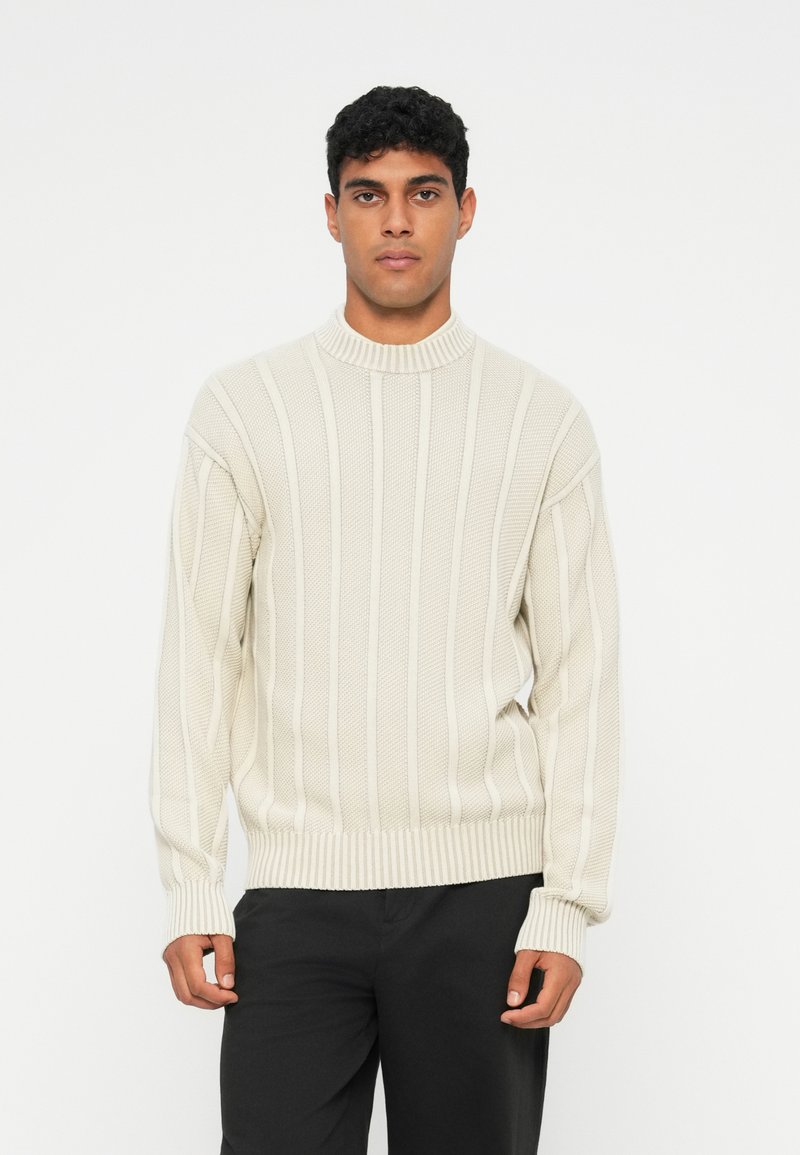 Pullover de malha em bege claro, com riscas verticais e um pescoço alto, apresentando um padrão texturizado e punhos e barra canelados.